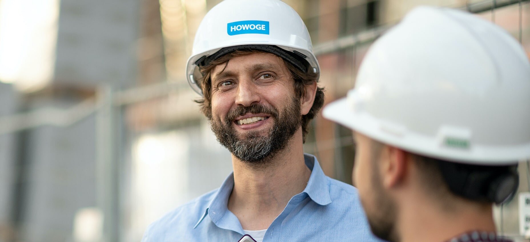 Ein lächelnder Mann mit weißem HOWOGE-Bauhelm schaut zu einem weiteren Mann mit Bauhelm, der von hinten zu sehen ist; im Hintergrund eine Baustelle.