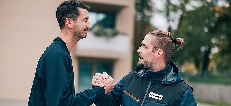 Zwei Männer begrüßen sich lächelnd mit Handschlag vor einem Wohngebäude.