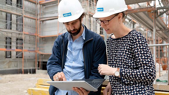Zwei Mitarbeiter mit Bauhelm schauen aufs Tablet, Sie stehen auf eine Baustelle