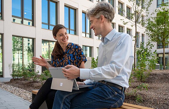 Zwei Menschen sitzen auf einer Bank vor der HOWOGE Zentrale. Im Hintergrund ein grüner Baum.