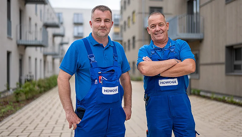 Zwei HOWOGE-Handwerker in blauer Arbeitskleidung stehen lächelnd zwischen Wohngebäuden.