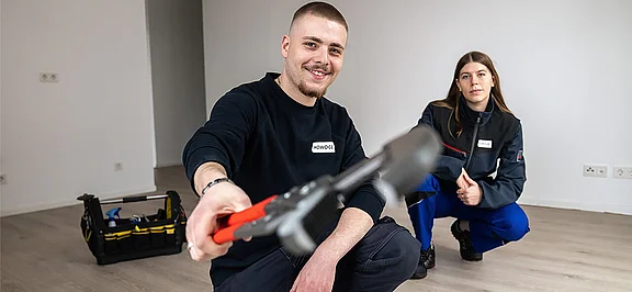 Zwei HOWOGE-Auszubildende in Arbeitskleidung knien in einer leeren Wohnung, der junge Mann hält lächelnd ein Werkzeug in die Kamera.