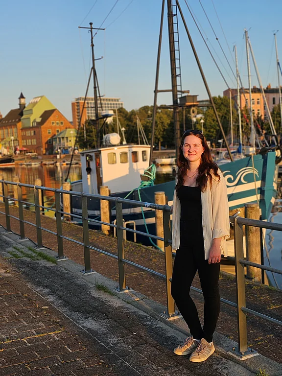 Eine Frau steht lachend auf einem Steg an einem Hafen, im Hintergrund ein Segelschiff.