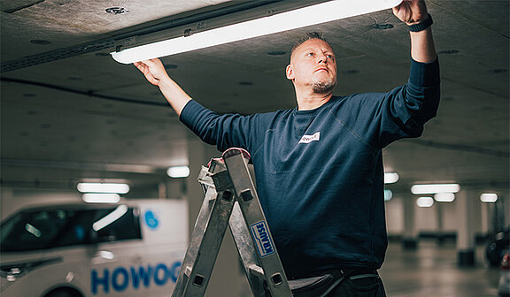 Ein Mitarbeiter der ATOZ Facility Solutions GmbH wechselt eine Lampe aus und steht dabei auf einer Leiter.