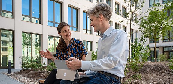zwei Menschen sitzen auf einer Bank, im Hintergrund die HOWOGE Zentrale. Man sieht grüne Bäume.