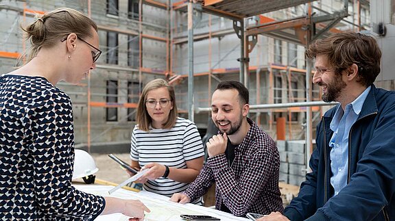 Vier Menschen stehen vor einem Bauplan auf der Baustelle