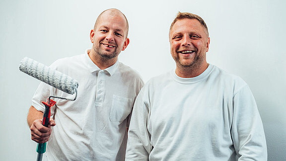 Foto von zwei freundlich lächelnden Malern, die in die Kamera schauen.