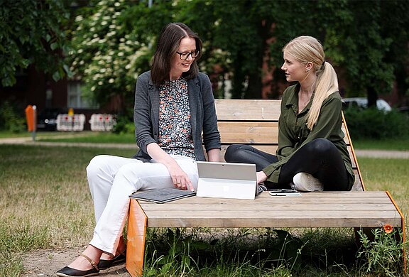 Werkstudentin sitzt mit ihrer Mentoring auf einer Bank. Sie unterhalten sich.
