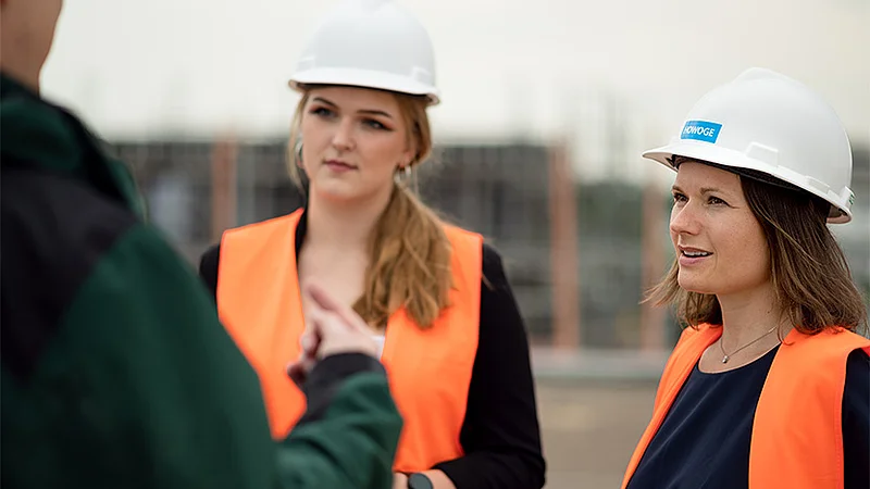 HOWOGE-Mitarbeiter:innen mit Bauhelm und Sicherheitsweste im technischen Austausch auf einer Baustelle.