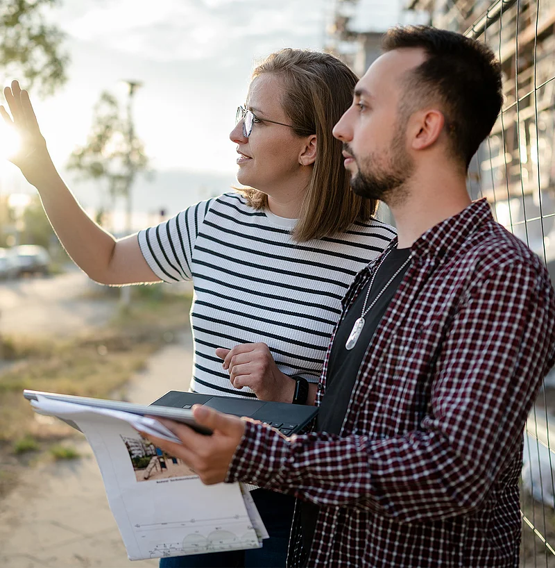 Eine Frau zeigt einem Studierenden an einer Baustelle auf ein Gebäude, während er einen Bauplan und Laptop in der Hand hält.