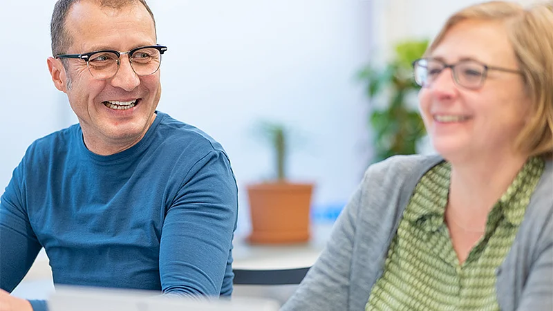 Zwei HOWOGE-Mitarbeitende lachen gemeinsam im Büro.