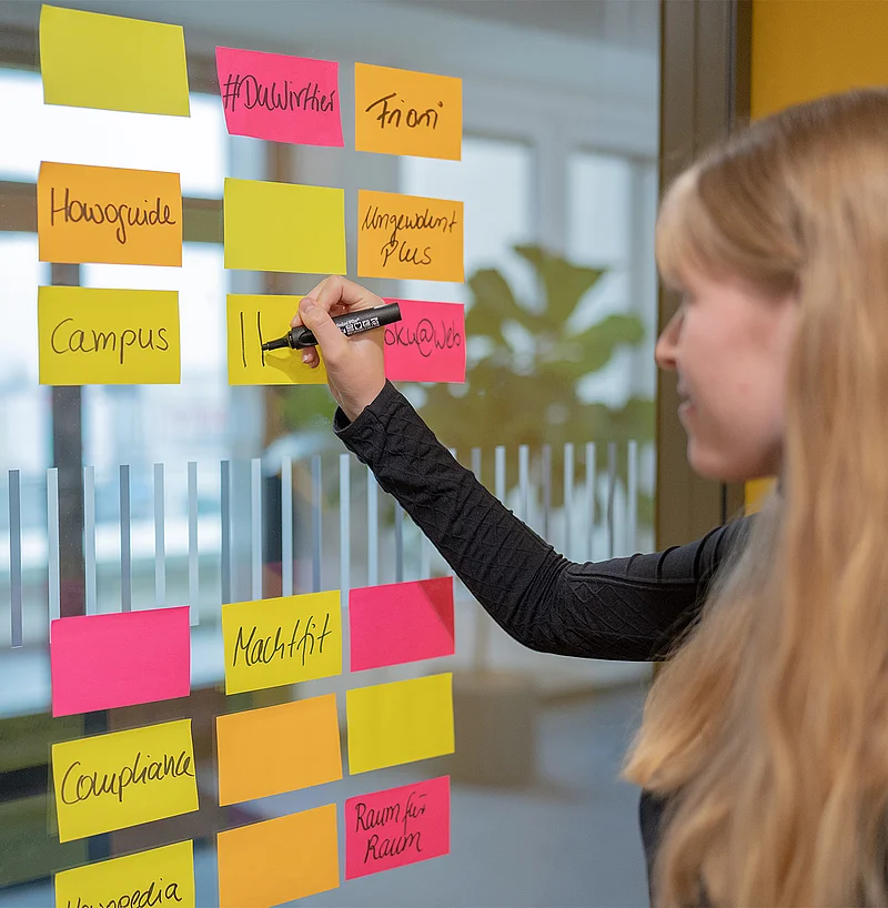 Eine Frau beschriftet bunte Klebezettel mit Projektbegriffen an einer Glaswand im Büro.