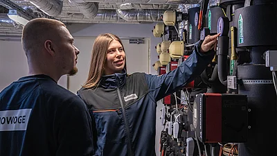 Eine Frau in HOWOGE-Arbeitsjacke zeigt lächelnd auf ein Anzeigeinstrument der Heizungsanlage, während ein Mann daneben zusieht.