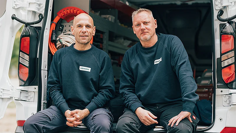 Zwei Handwerker in HOWOGE-Arbeitskleidung sitzen lächelnd an der offenen Ladefläche eines Firmenfahrzeugs.