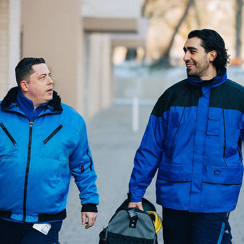 Zwei Männer in blauen Jacken schauen sich lächelnd an und gehen mit einer Werkzeugtasche eine Straße entlang.