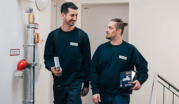 Zwei MItarbeiter der ATOZ Facility Solutions GmbH laufen einen Flur entlang und unterhalten sich. Dabei schauen sie sich an und lächeln.