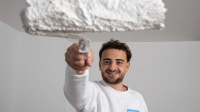 Lächelnder junger Mann hält einen Farbroller mit weißer Farbe in die Kamera.