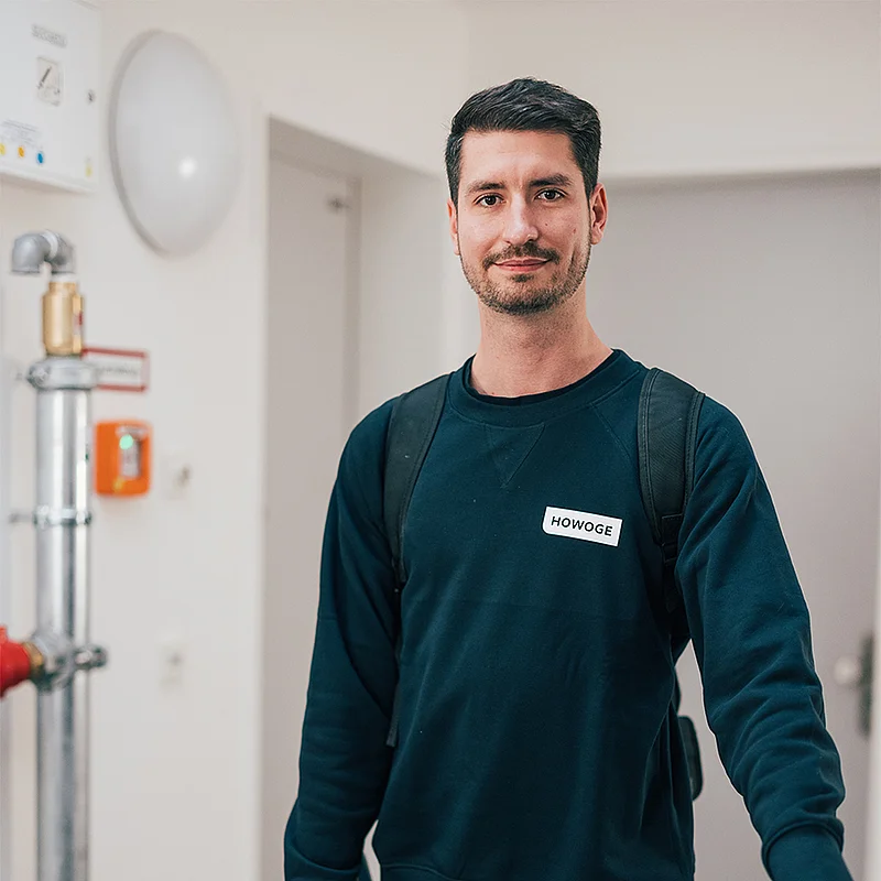 Max Strey, Vorarbeiter HLS-Reparaturen, steht mit Rucksack in dunklem HOWOGE-Pullover in einem Flur.