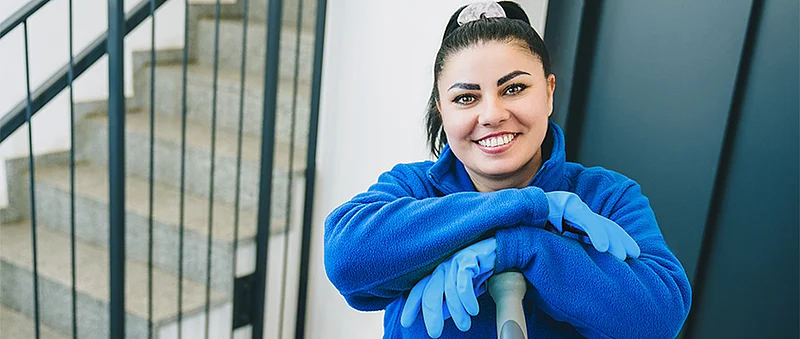 Eine lächelnde Frau in blauer Arbeitskleidung und blauen Handschuhen lehnt sich auf ein stielartiges Reinigungsgerät, neben ihr ist ein Treppengeländer zu sehen.