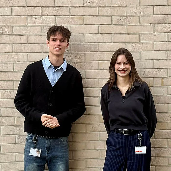 Ein junger Mann und eine junge Frau stehen lächelnd nebeneinander vor einer hellen Backsteinwand.