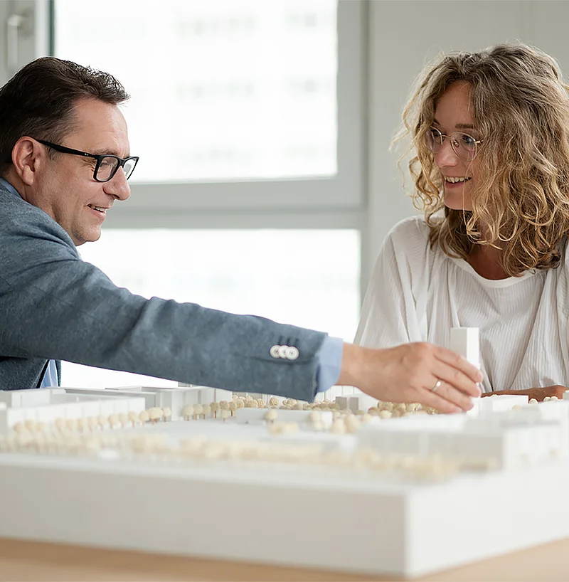 Mann und Frau besprechen lächelnd ein Architekturmodell einer Wohnsiedlung.