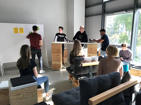 Mehrere Personen sitzen und stehen in einem Schulungsraum an Tischen und einem Whiteboard.