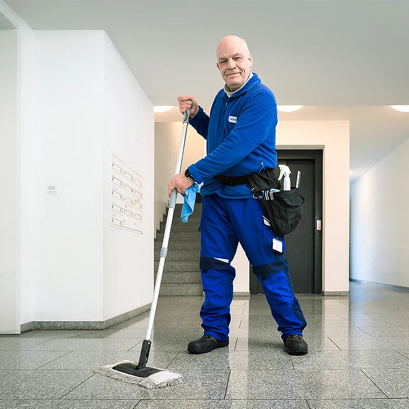 Andreas Feddermann; Reinigungskraft, steht mit einem Wischmopp in blauer HOWOGE-Arbeitskleidung in einem Hausflur.