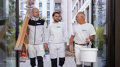 Drei Männer in weißer Malerkleidung gehen mit Werkzeug und Eimern draußen vor einem Gebäude.