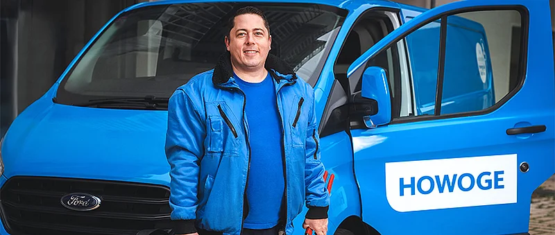 Björn Biermann steht lachend in blauer Jacke vor einem blauen HOWOGE-Firmenwagen.