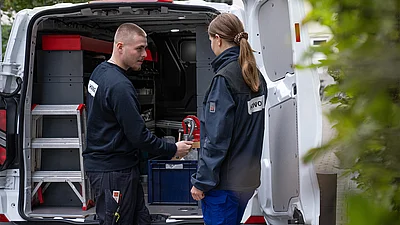 Ein Mann und eine Frau in dunkler Arbeitskleidung stehen an der geöffneten Heckklappe eines Firmentransporters und prüfen Werkzeug/Ausrüstung.