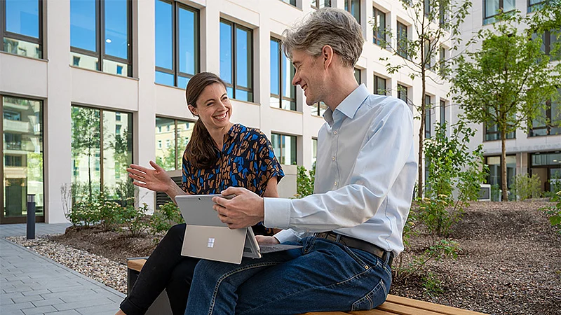Zwei HOWOGE-Mitarbeitende sitzen lachend mit einem Laptop auf einer Bank vor dem Bürogebäude.