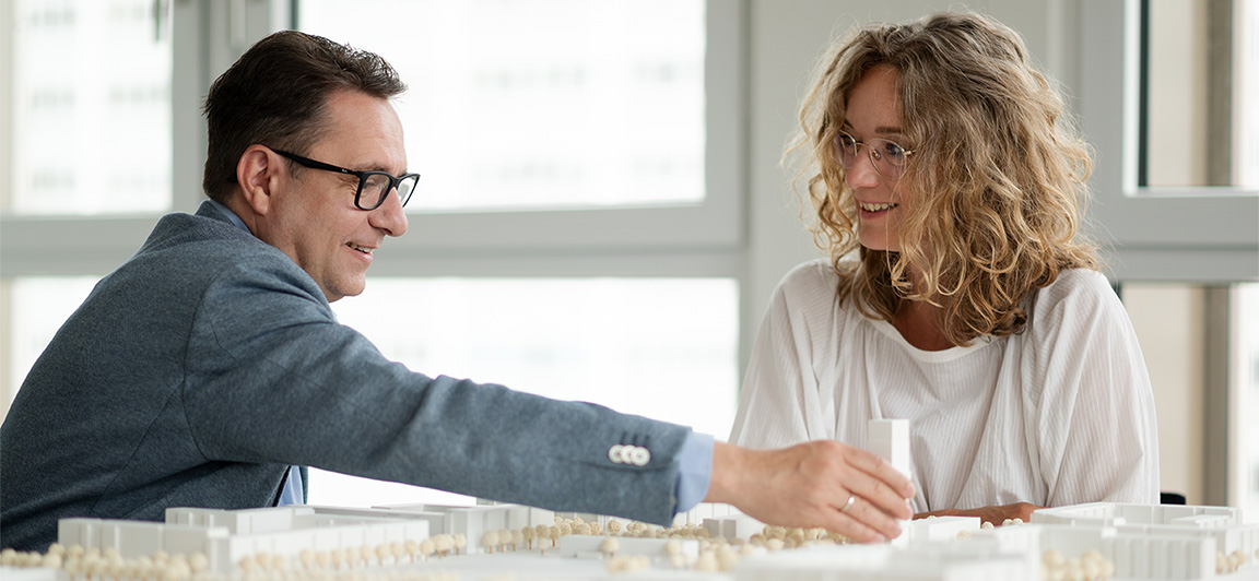 Mann und Frau besprechen lächelnd ein Architekturmodell einer Wohnsiedlung.
