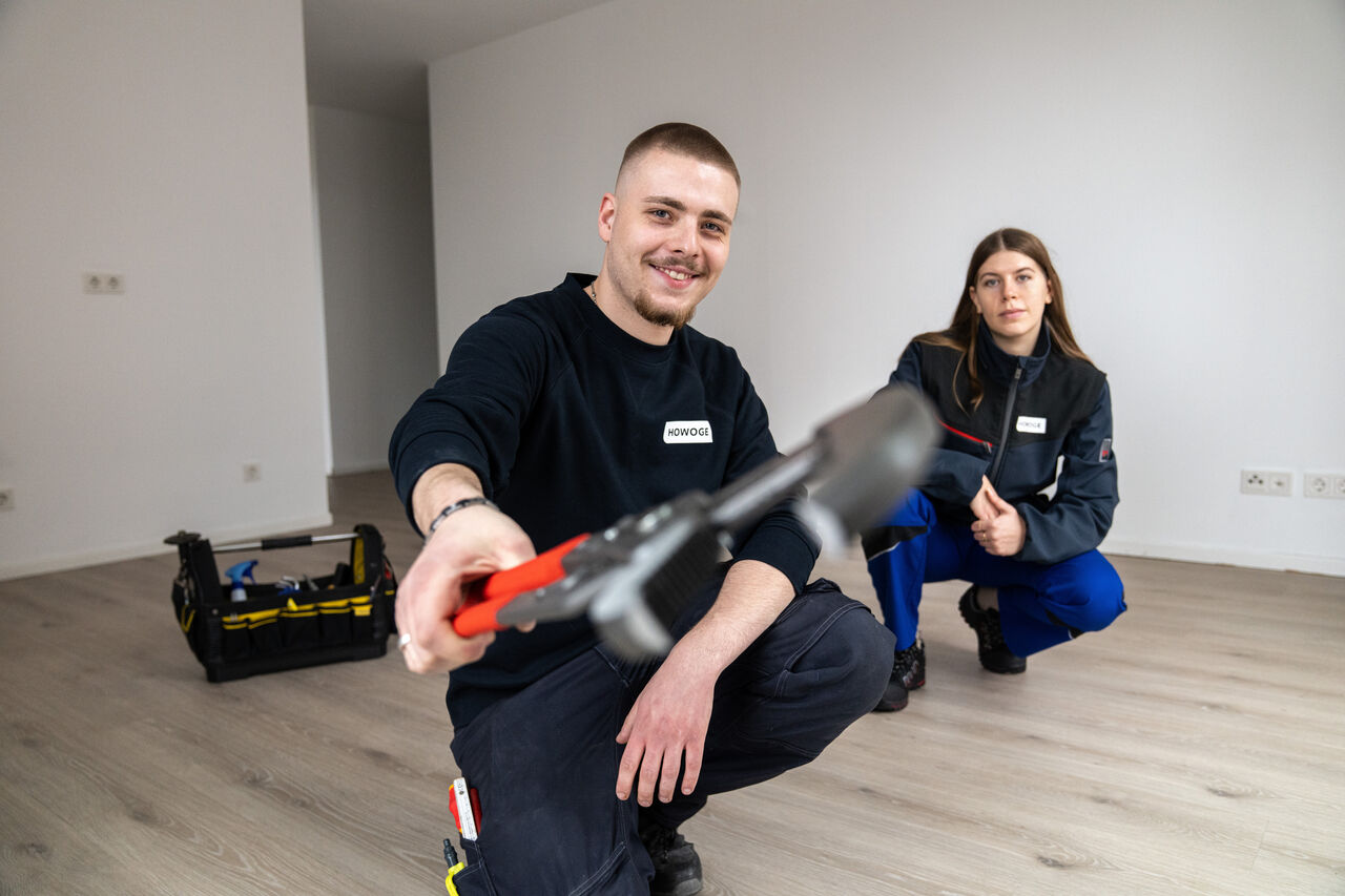 Zwei HOWOGE-Auszubildende in Arbeitskleidung knien in einer leeren Wohnung, der junge Mann hält lächelnd ein Werkzeug in die Kamera.