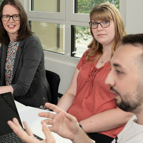 Drei Personen sitzen an einem Tisch mit Laptop und Unterlagen und diskutieren miteinander, ein Mann im Vordergrund gestikuliert beim Sprechen.